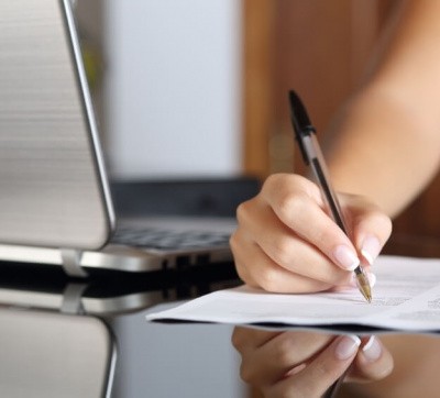Close up of a woman hand writing a contract with a laptop beside at home or office